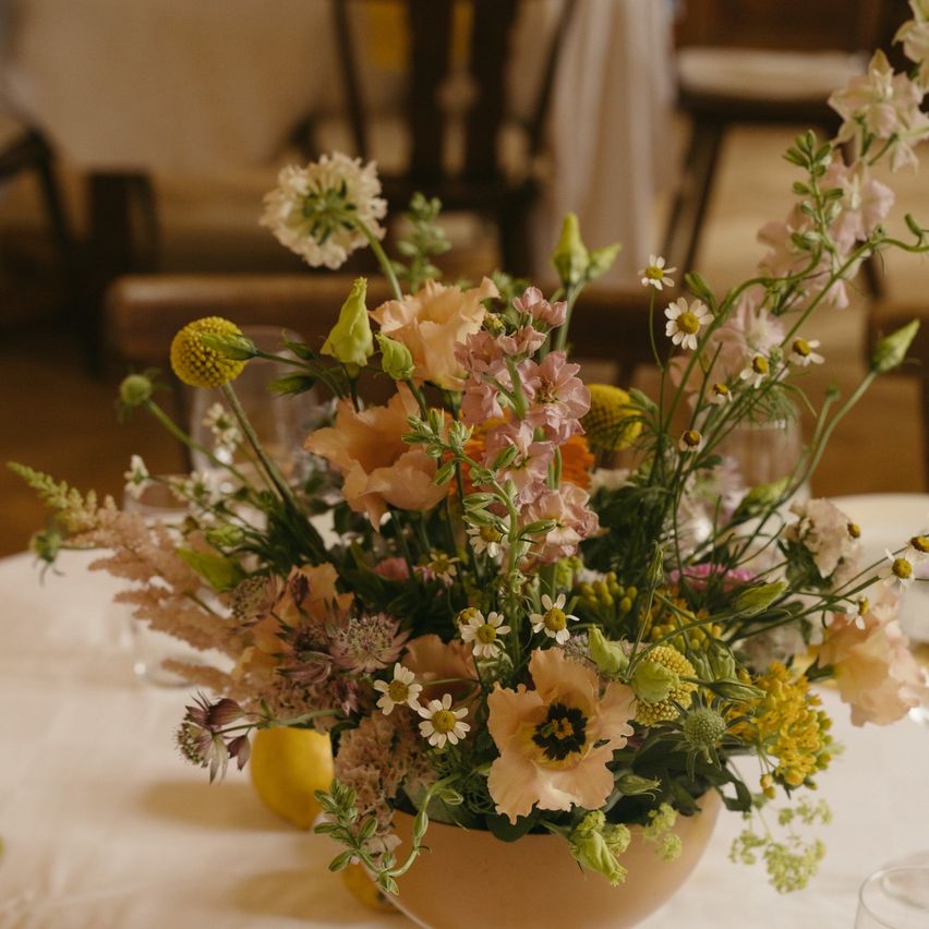 Ein Tisch mit einer Blumendekoration in einem beigefarbenen Gefäß. Die Blumen sind eine Mischung aus Weiß, Rosa und Gelb.