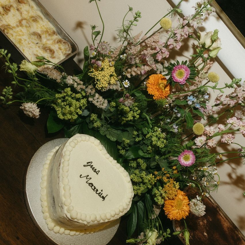 Ein herzförmiger Kuchen mit den Worten 'Just Married' darauf steht auf einem Teller. Ein Strauß bunter Blumen daneben auf einem Holztisch. Ein Tablett mit Essen ist hinter dem Strauß.