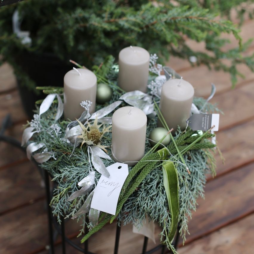 Ein kreisförmiger Kranz aus grünem Laub und silbernen Bändern umgibt fünf Kerzen, verziert mit dekorativen Ornamenten, auf einer Holzoberfläche.