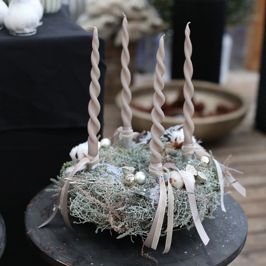 Ein dekorativer Kranz mit verdrehten Kerzen und weißem Moos schmückt einen Holztisch. Ein mit Tannenzapfen gefüllter Korb steht im Hintergrund.