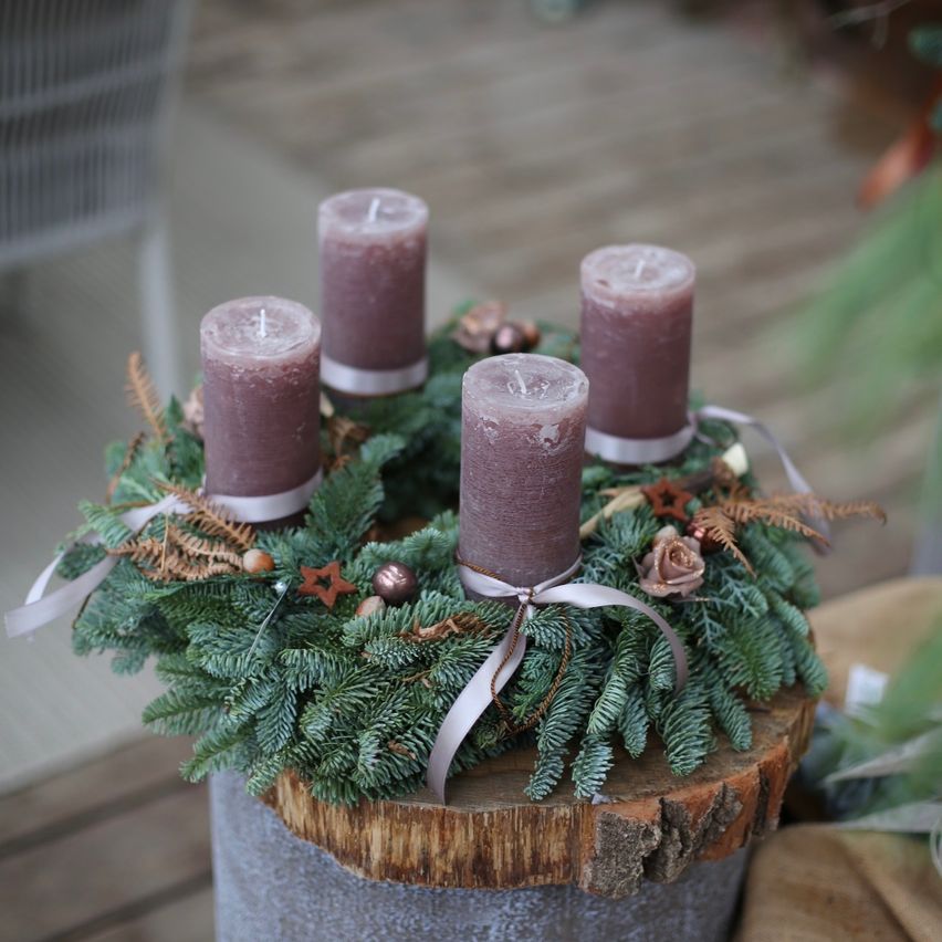 Ein Kranz mit vier lavendelfarbenen Kerzen, geschmückt mit Tannenzweigen, Sternen und einem Band, liegt auf einem Baumstumpf.