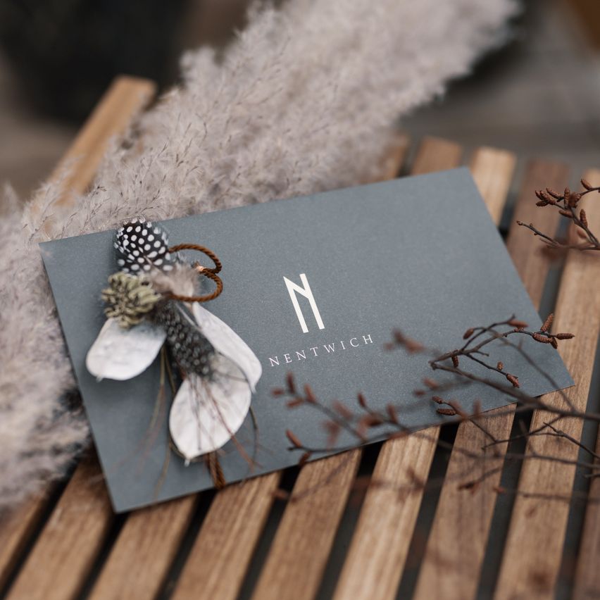 Eine graue Visitenkarte mit dem Logo von NENTWICH liegt auf einer Holzoberfläche, verziert mit einer weißen Feder und grauem Fell.