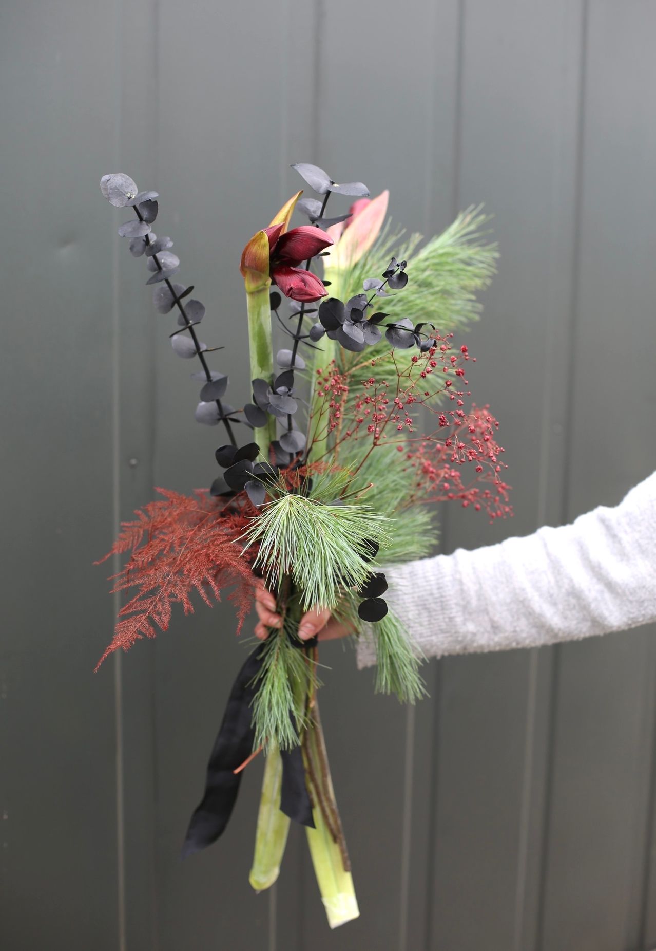 Eine Nahaufnahme eines Blumenstraußes mit dunkelroten Blättern und schwarzen Stielen, mit einer auffälligen roten Blüte und grünem Laub, gehalten von einer Person in einem weißen Langarmshirt.