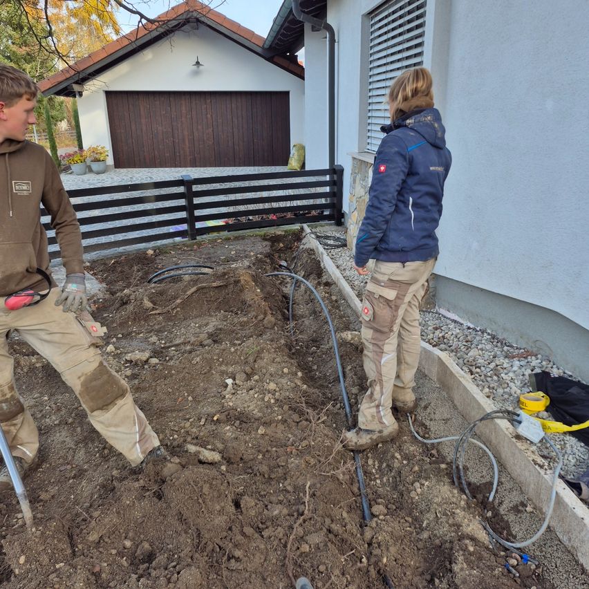 Zwei Personen arbeiten vor einem Haus. Einer gräbt, während der andere zuschaut. Sie tragen beide Jacken und Hosen. Ein Zaun und eine Garage sind hinter ihnen.