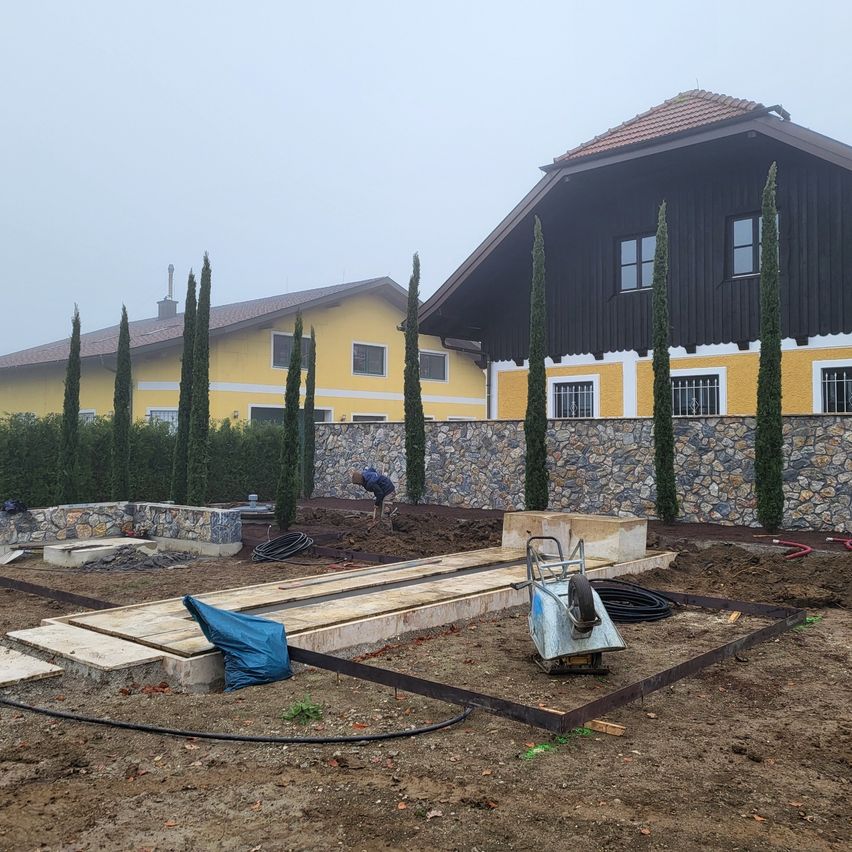 Eine Baustelle mit einem gelben Haus im Hintergrund. Eine Person arbeitet am Boden. Ein Schubkarren und eine blaue Plane sind in der Nähe.