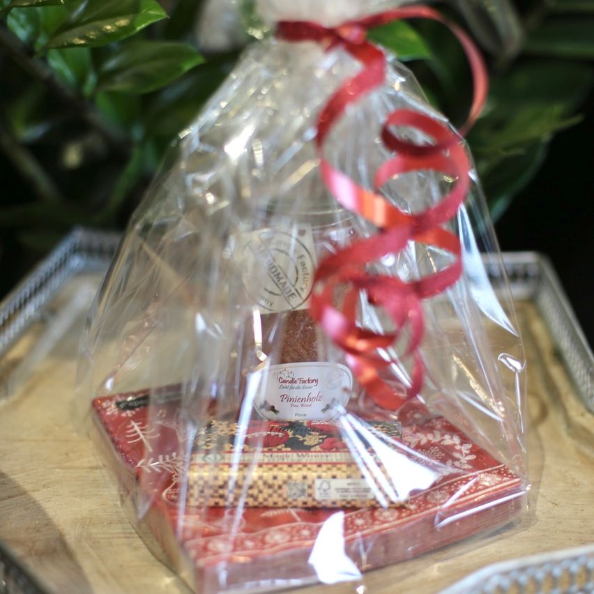 Ein durchsichtiger Plastikbeutel mit einem roten Band enthält ein Geschenkset, bestehend aus einer Kerze und einer kleinen Schachtel, auf einem rot gemusterten Karton. Die Kerze hat ein Etikett mit der Aufschrift 'Kerzenfabrik'. Hinter dem Beutel befinden sich grüne Blätter.