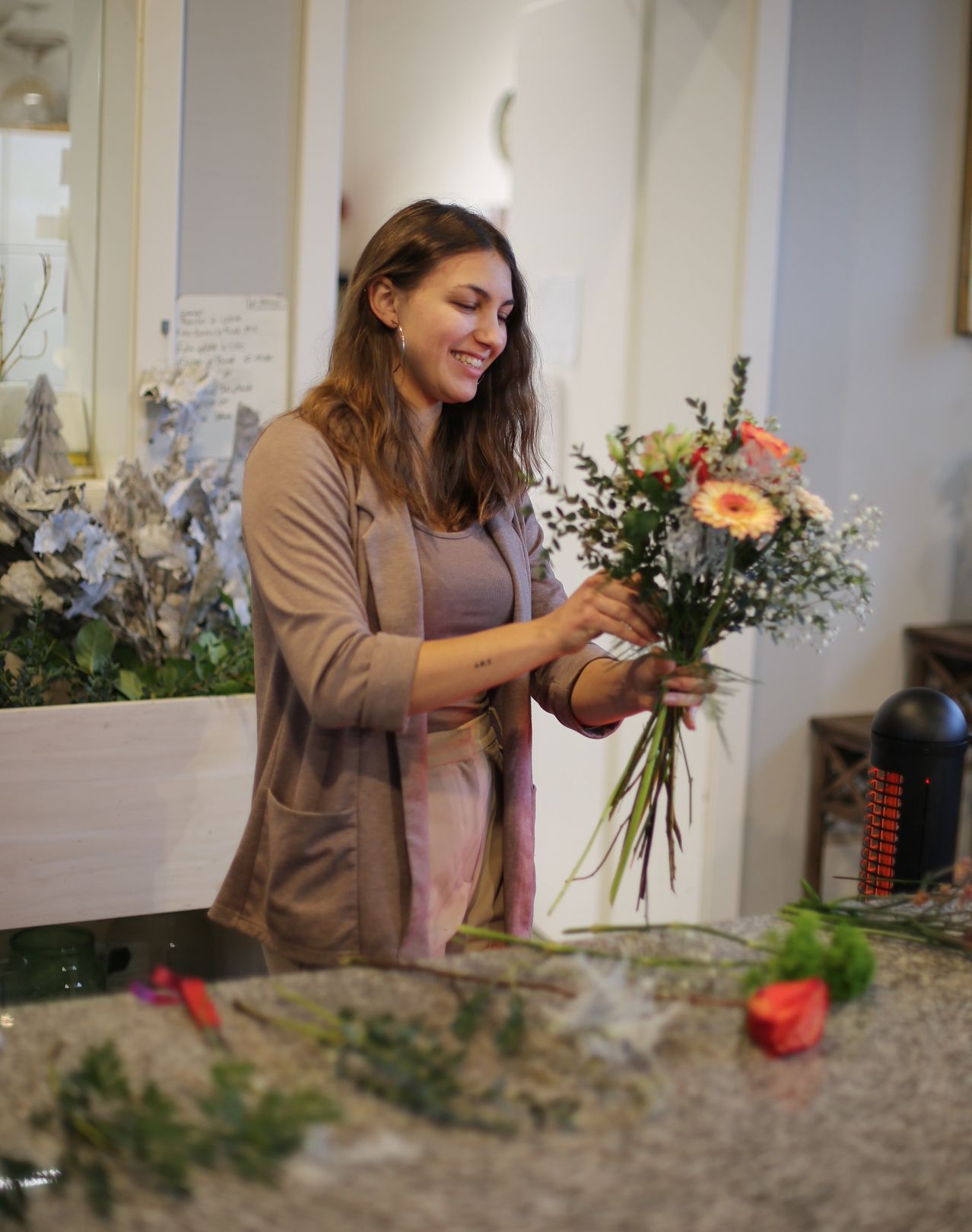 Eine Frau arrangiert einen Blumenstrauß auf einem Tisch. Sie trägt eine braune Strickjacke und einen rosa Rock. Der Strauß enthält Rosen, Gänseblümchen und Grün. Ein Mülleimer ist in der Nähe.