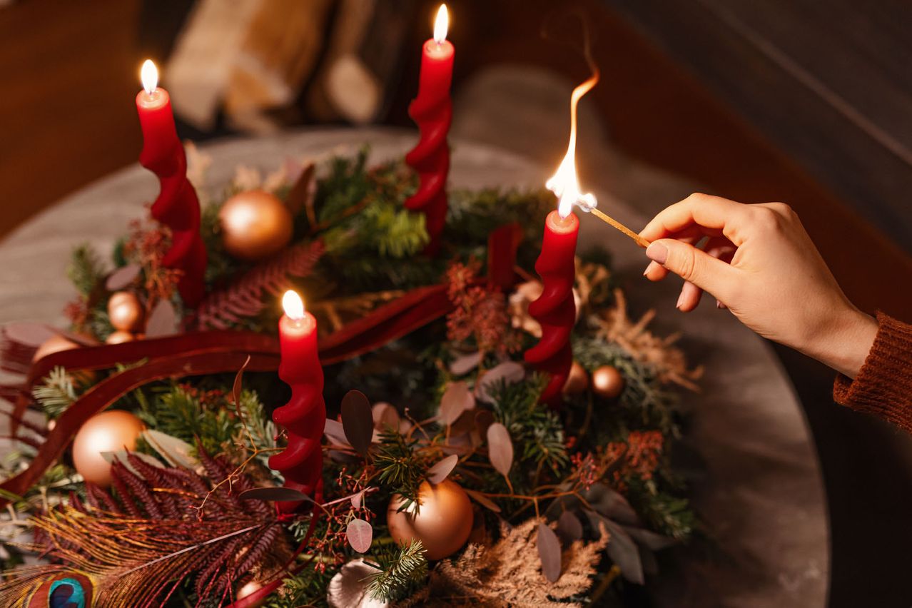 Eine Hand zündet eine Kerze mit einem Streichholz an. Die Kerze ist rot und hat eine Spiralform. Die Kerze steht auf einem Adventskranz. Der Adventskranz hat ein rotes Band und eine rote Kugel. Der Adventskranz ist mit grünen Blättern und getrockneten Blumen verziert.