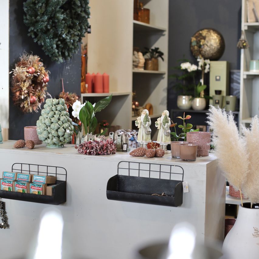 Ein Raum gefüllt mit Dekorationsartikeln, darunter ein weißer Tresen mit verschiedenen Pflanzen, Figuren und Kerzen. Topfpflanzen, ein Kranz und Regale mit verschiedenen Gegenständen sind ebenfalls vorhanden.