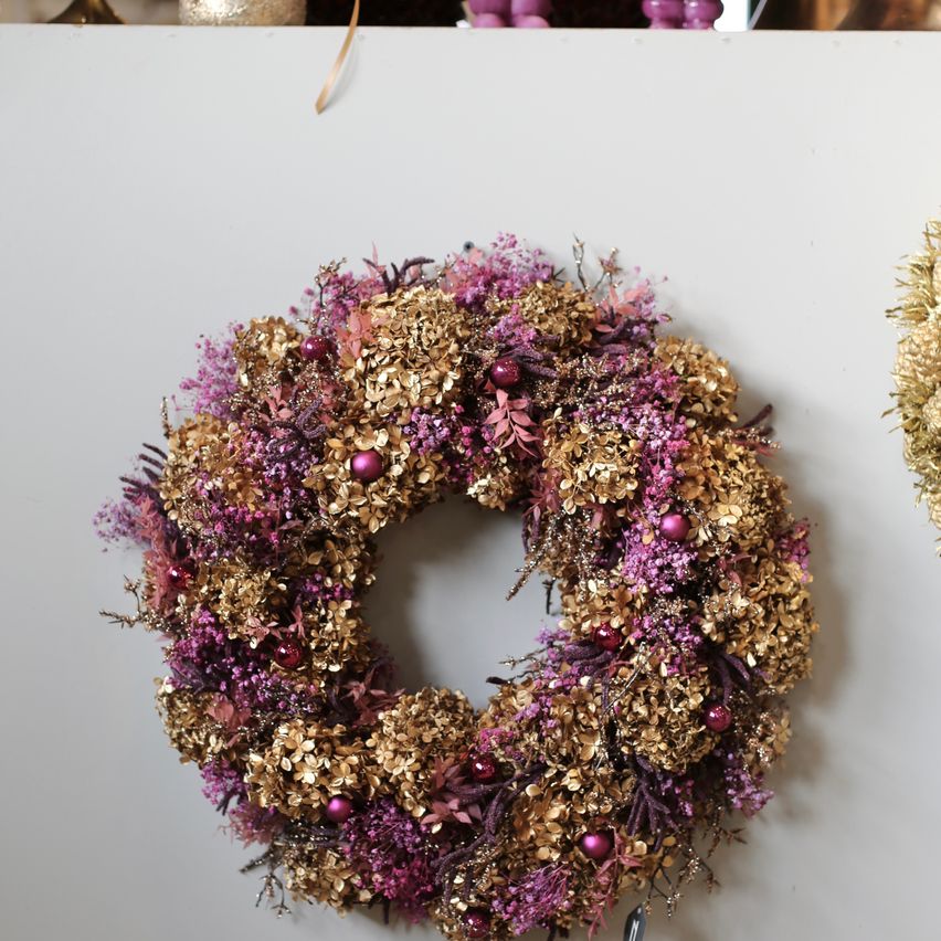 Ein dekorativer Kranz mit goldenen und lila Blumen und roten Ornamenten hängt an einer weißen Wand.