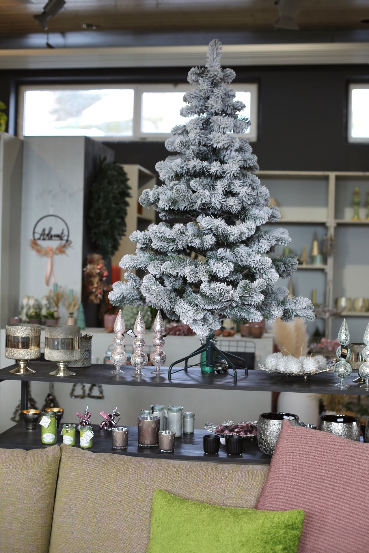 Ein weißer, frostbedeckter künstlicher Weihnachtsbaum steht prominent in einem Geschäft, umgeben von verschiedenen Dekorationen, darunter silberne Ornamente, Glasschalen und eine rosa Kissen auf einem nahegelegenen Sofa.