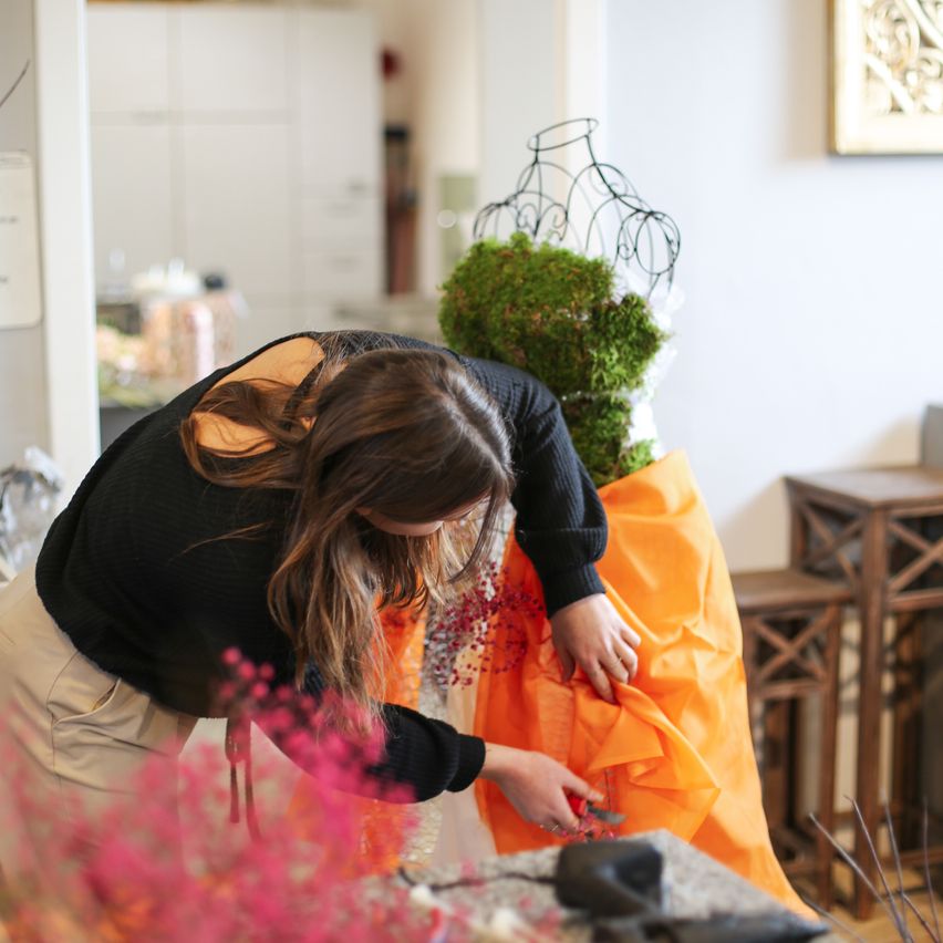 Eine Frau arbeitet an einem orangefarbenen Kleid, das mit Moos bedeckt ist, eine Schere in der Hand, umgeben von rosa Blumen und einem Metallmännchen auf einem Tisch.