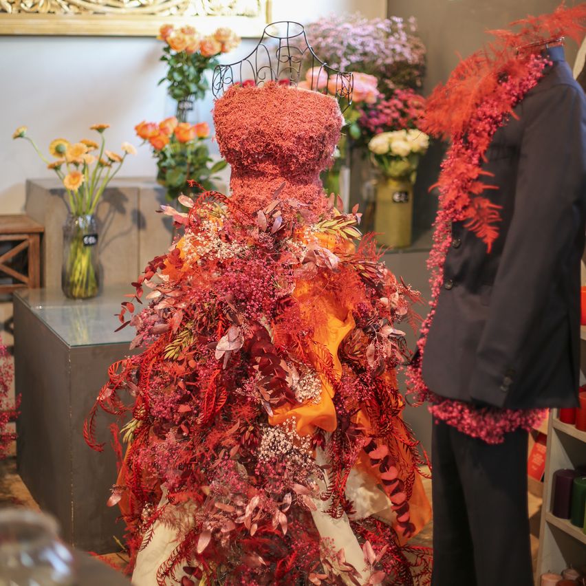 Ein leuchtendes rotes und orangefarbenes Blumenkleid mit Blättern wird auf einem Mannequin ausgestellt. Ein dunkler Mantel und eine Krawatte sind in der Nähe. Der Hintergrund besteht aus weißen Wänden und einer Vielzahl von Blumen in Vasen.