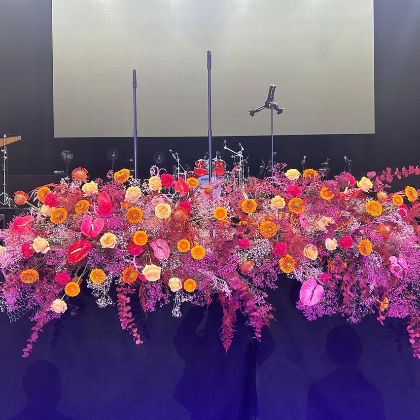 Eine farbenfrohe Blumendekoration auf einer Bühne, bestehend aus einer Mischung aus roten, rosa, gelben und orangefarbenen Blumen, mit Mikrofonen und Musikinstrumenten im Hintergrund.