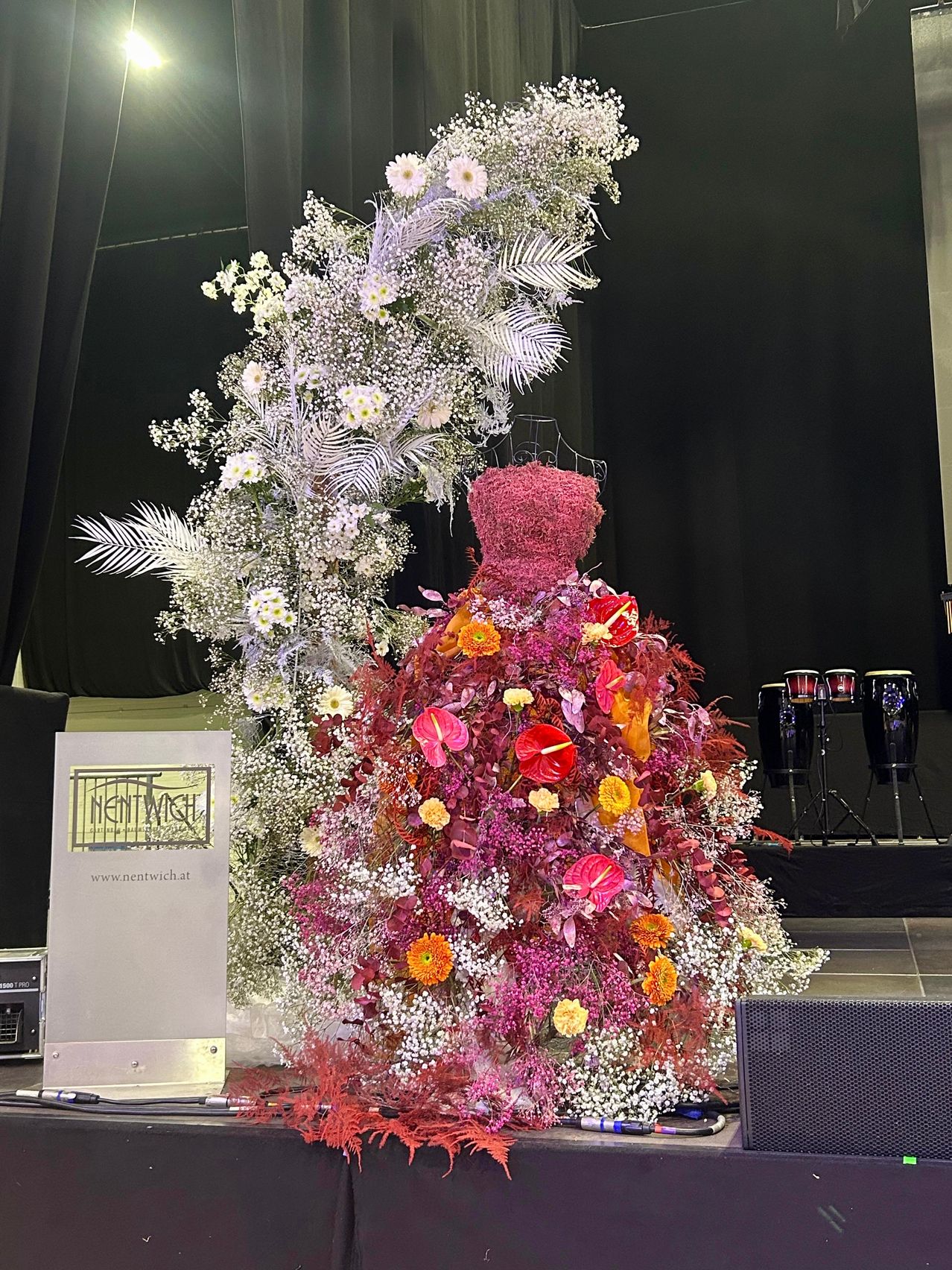 Ein Blumenkleid wird auf einem Mannequin vor einem schwarzen Vorhang ausgestellt. Links befindet sich ein Schild für Nentwich. Hinter dem Kleid stehen drei Bongo-Trommeln auf Ständern.