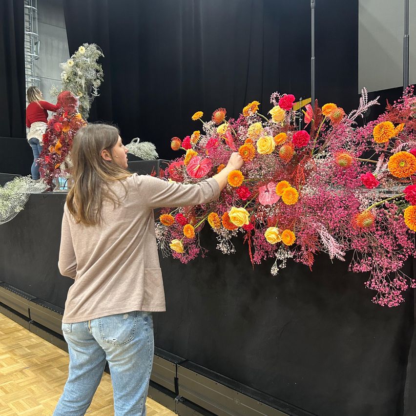 Eine Frau steht vor einer Blumendekoration an einer schwarzen Wand und betrachtet sie. Im Hintergrund arbeitet eine andere Person an einer Blumenarrangement.