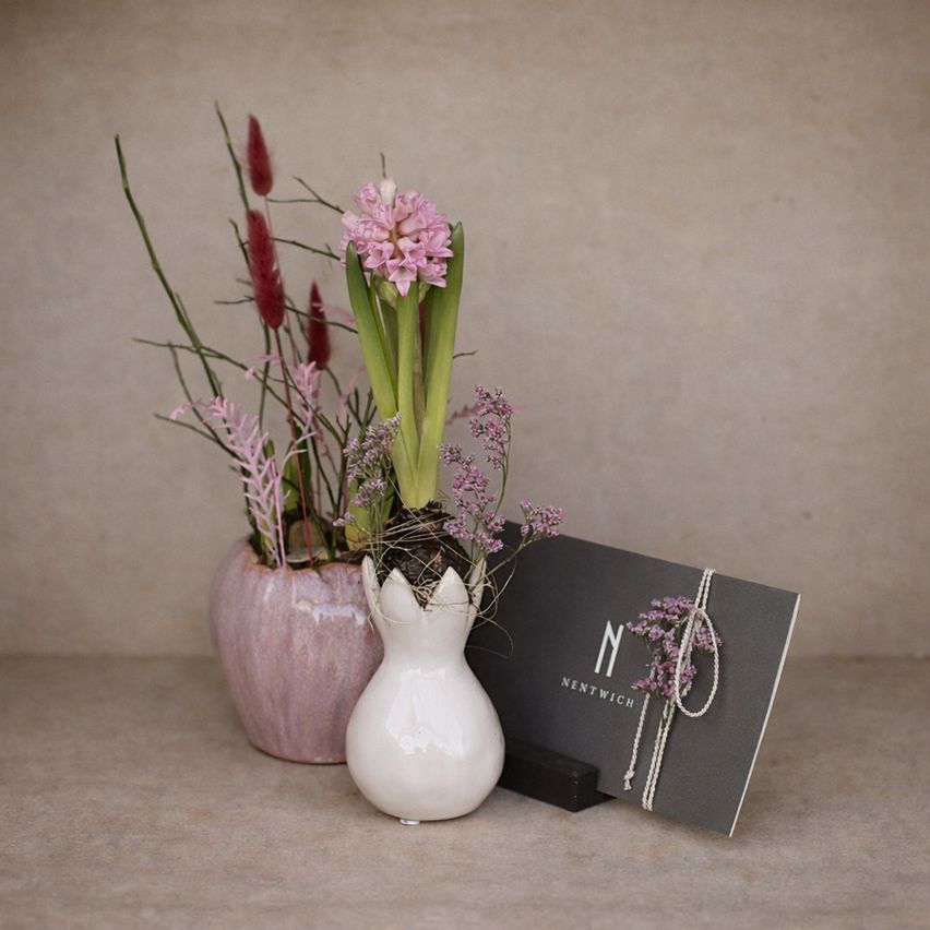 Zwei Vasen mit rosa Blumen stehen auf einem Tisch, eine mit einem rosa Topf und die andere mit einem weißen Topf. Ein Karten mit dem Text 'NENTWICH' steht neben der weißen Vase.