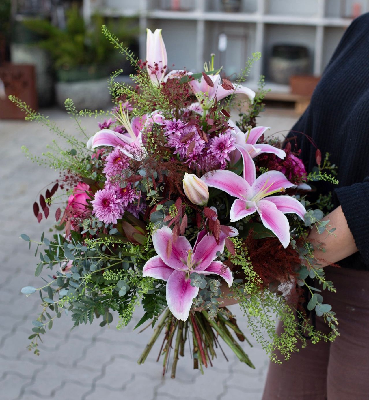 Eine Person hält einen Blumenstrauß aus rosa Lilien, lila Blumen und grünem Laub. Der Strauß ist ordentlich arrangiert und wird von den Händen einer Person gehalten.