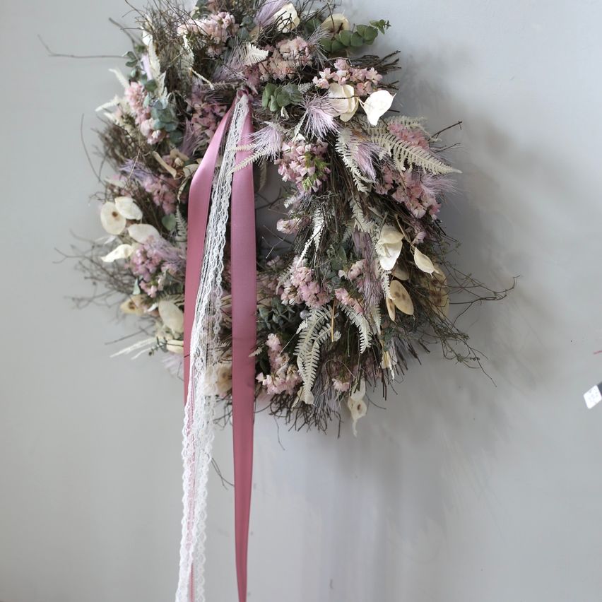 Ein kreisförmiger Kranz aus getrockneten Blumen und Grün mit einem rosa Band hängt an einer weißen Wand.