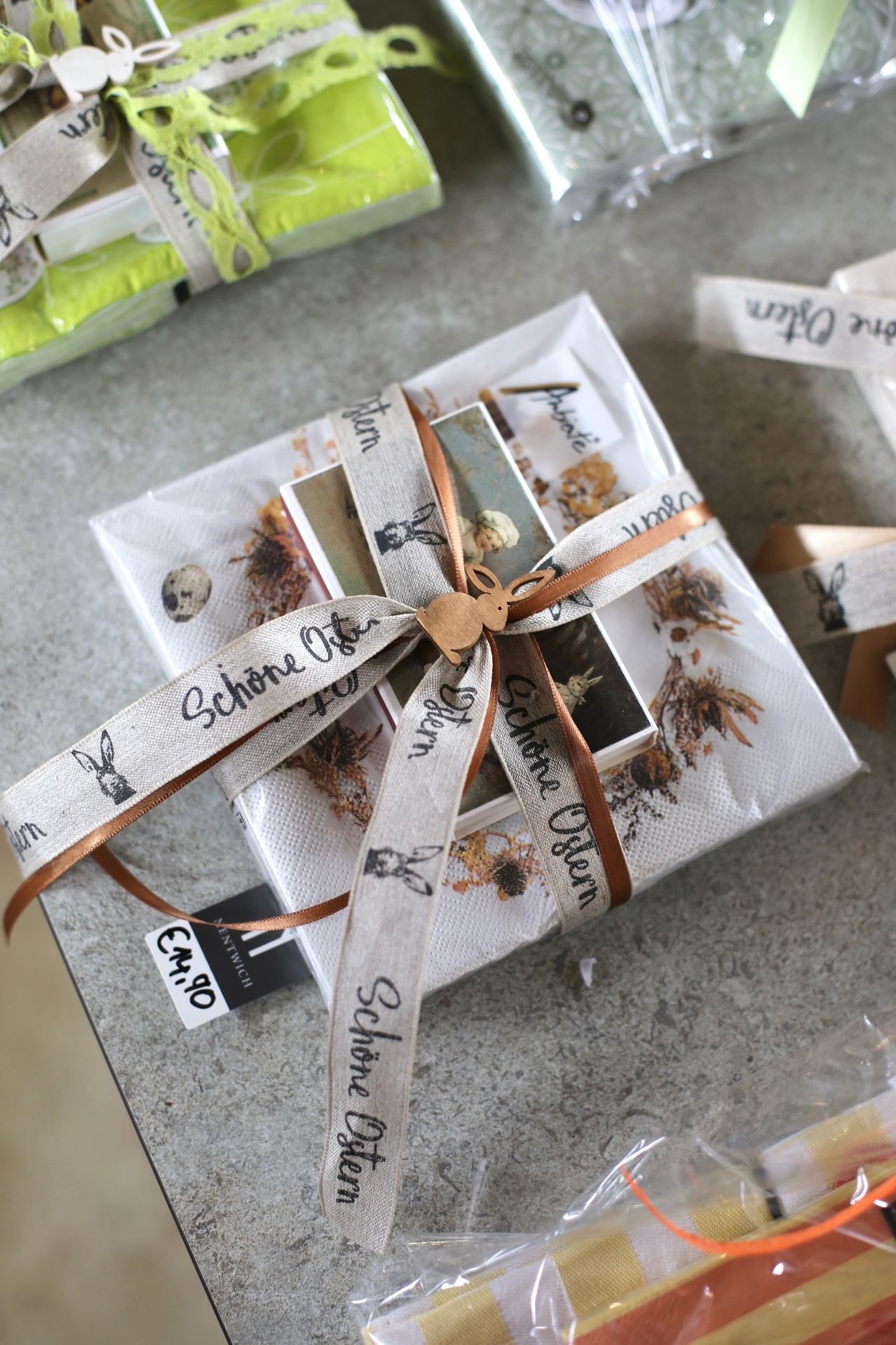 Ein verpacktes Geschenk mit einem braunen Band und einem Schild mit der Aufschrift 'Schone Ostern' liegt auf einer grauen Oberfläche. Die Verpackung enthält Ostereier und getrocknete Blumen.