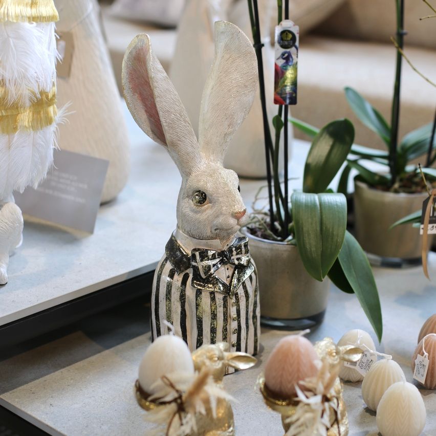 Eine dekorative Hasenfigur im Fliege, umgeben von Ostereiern und Pflanzen. Eine weiße Hasenstatue sticht unter anderen Dekorationsartikeln hervor.