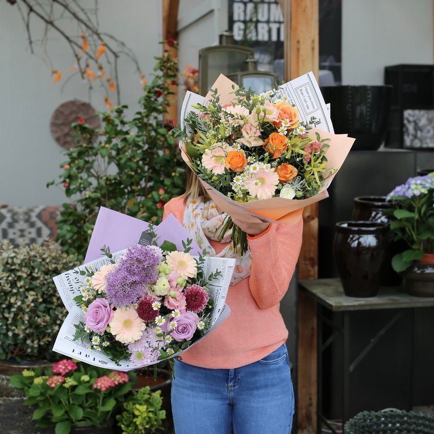 Eine Frau in einem rosa Pullover und blauer Jeans hält zwei Blumensträuße in einem Blumenladen.