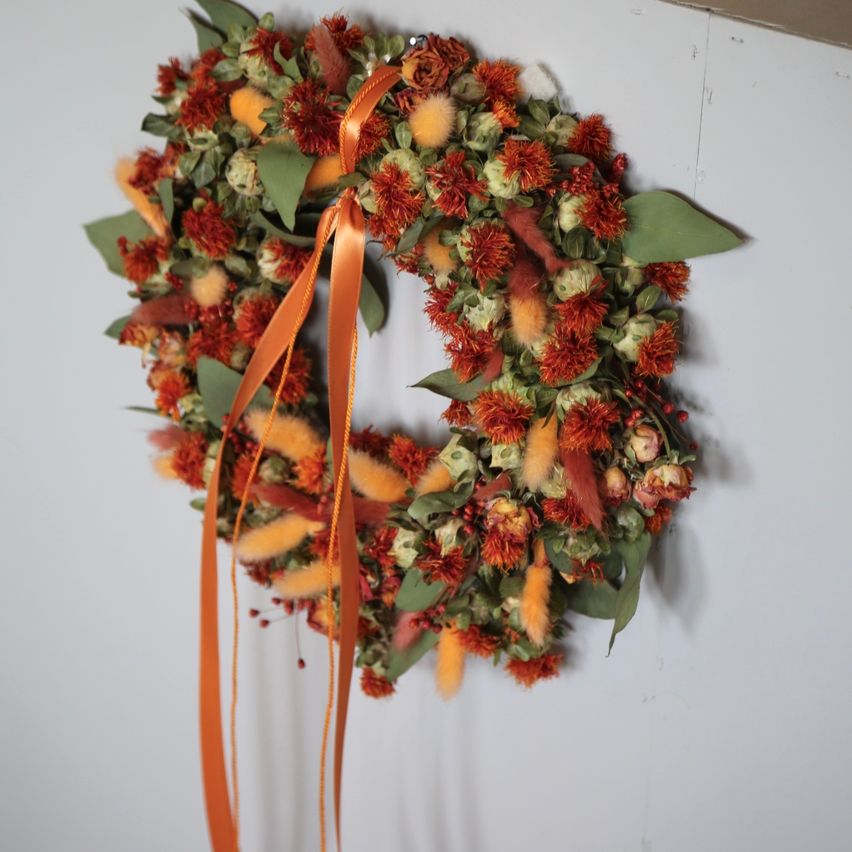 A wreath made of dried flowers and leaves, with an orange ribbon, hanging on a white wall.