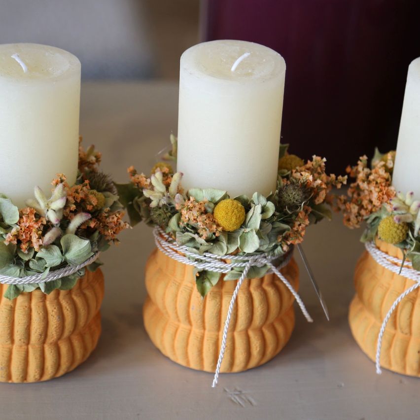 Three decorative candles with pumpkin-shaped holders, adorned with dried flowers and leaves, on a table.