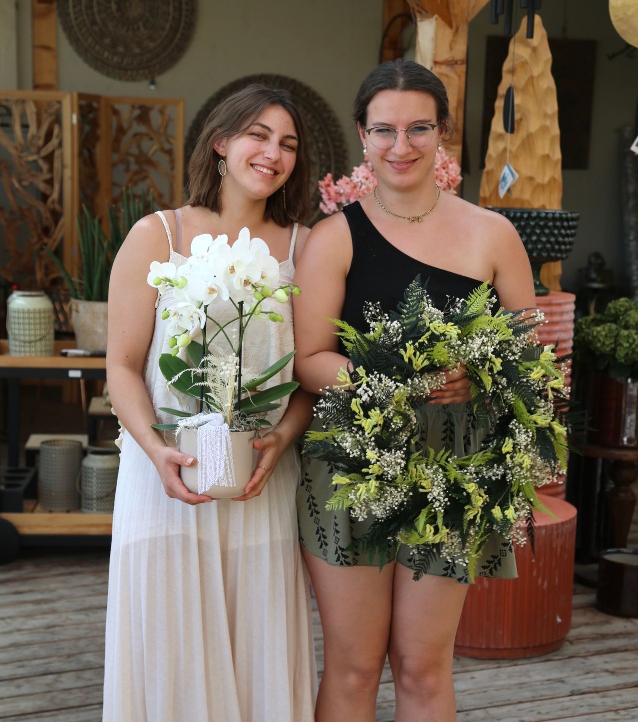Two women stand next to each other, one holding a white orchid in a pot and the other holding a green floral wreath. They are smiling and appear to be posing for a photo.