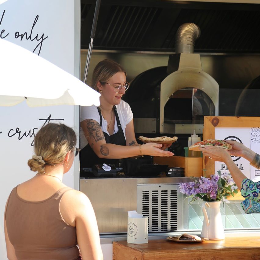 A woman in a white apron serves pizza through a food truck window. Customers receive their orders. An umbrella and a vase of flowers are nearby.