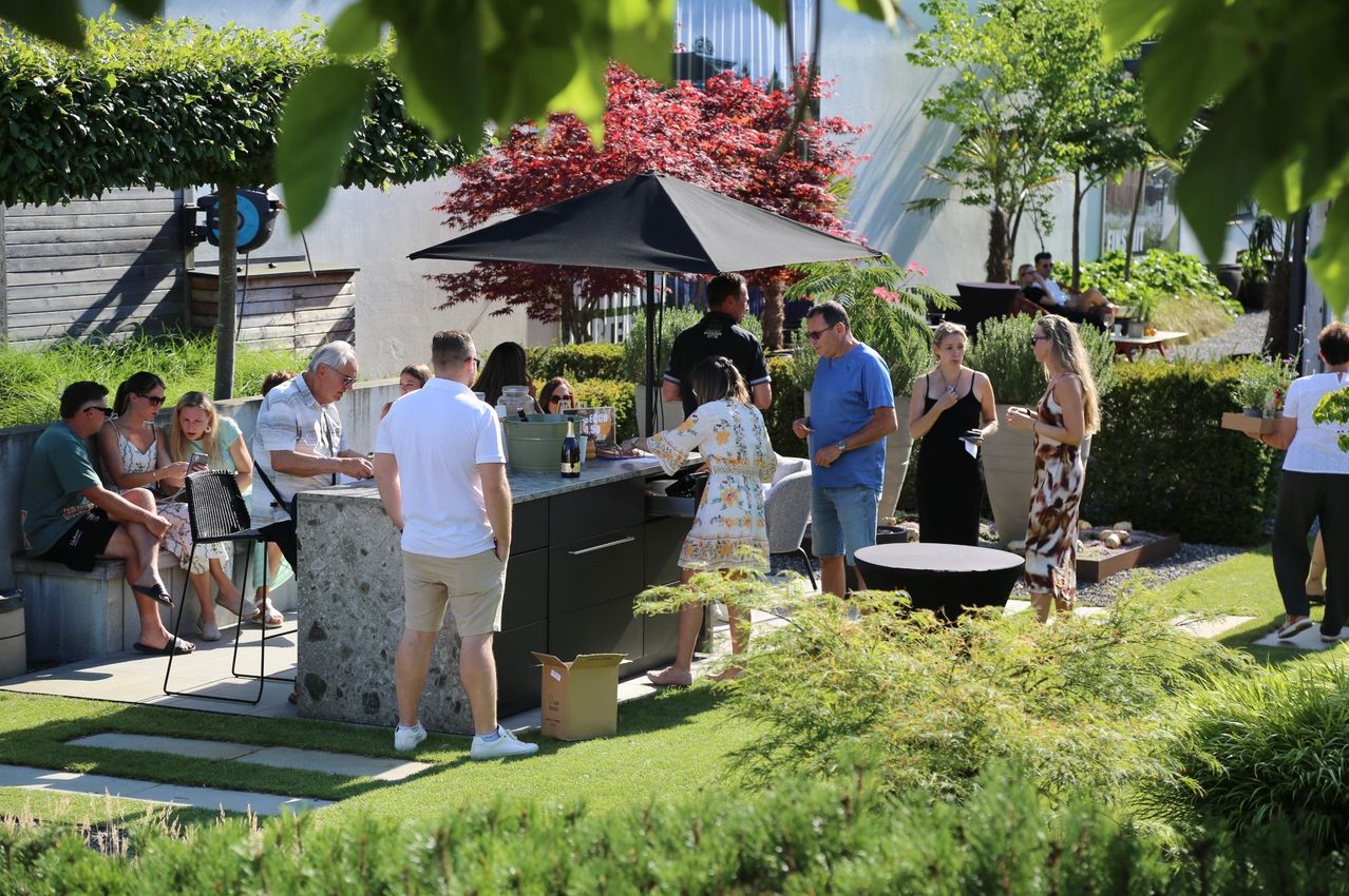 A group of people gather around a bar in a garden, some standing under a black umbrella, others chatting and enjoying the outdoor setting.