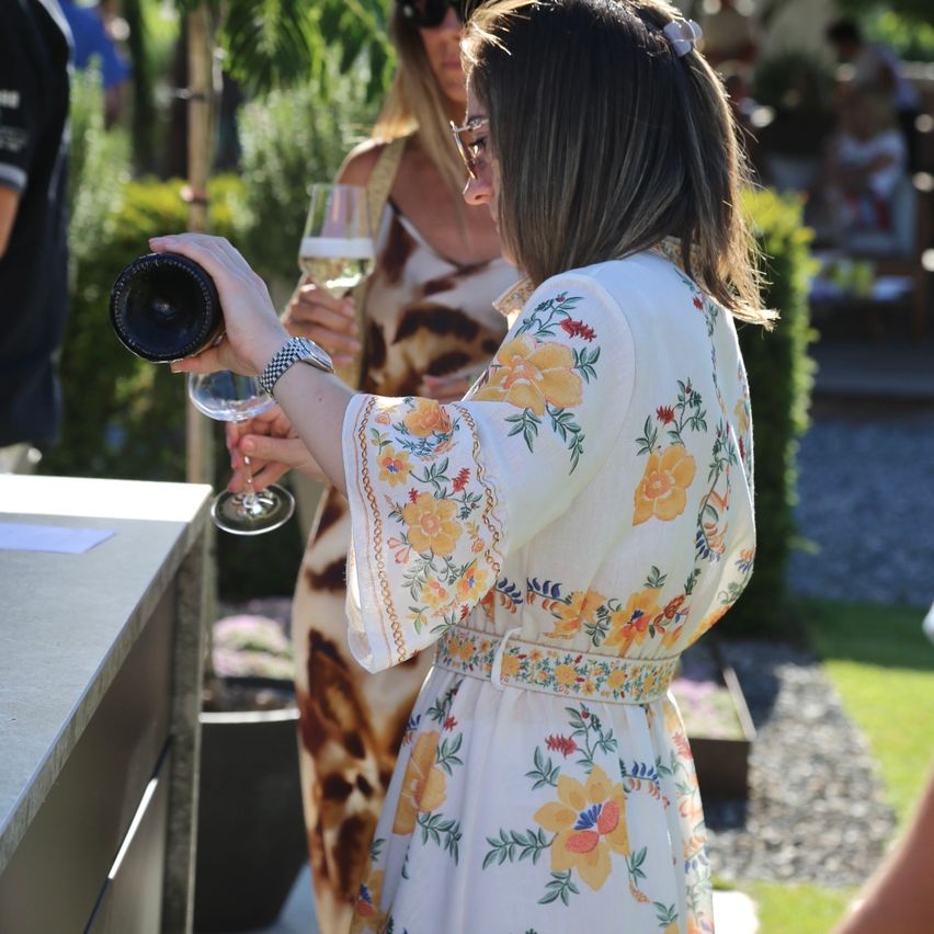 A woman opens a wine bottle while holding a wine glass, another woman watches. They stand in a garden.