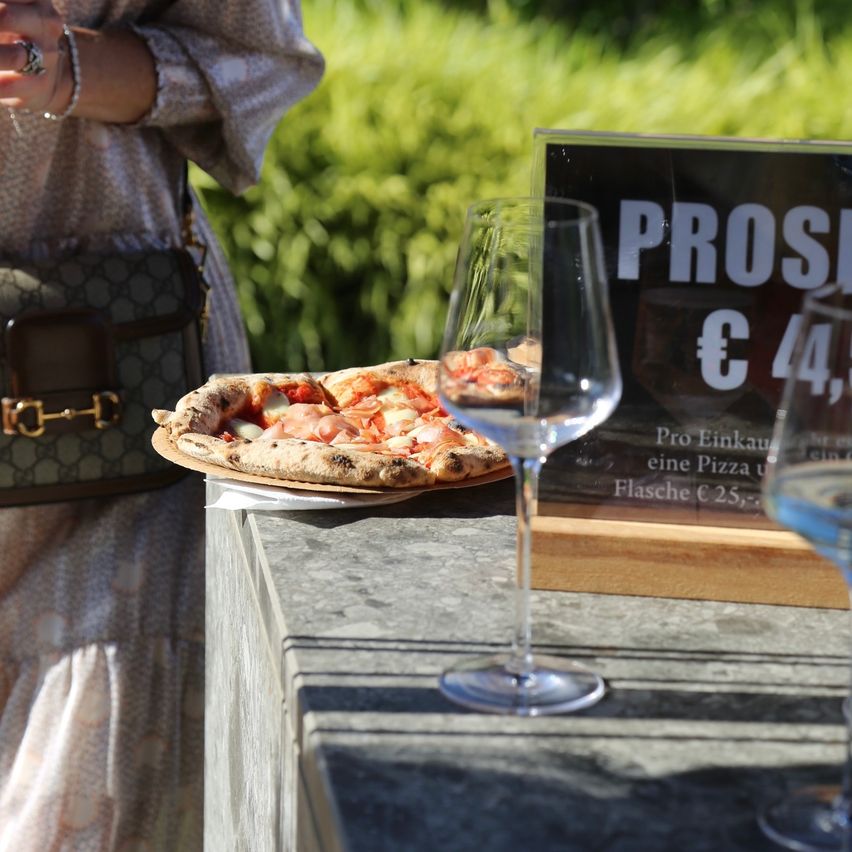 A pizza on a plate, with a wine glass next to it, and a signboard that reads PROS 4,5. A woman wearing a dress is standing beside the table.