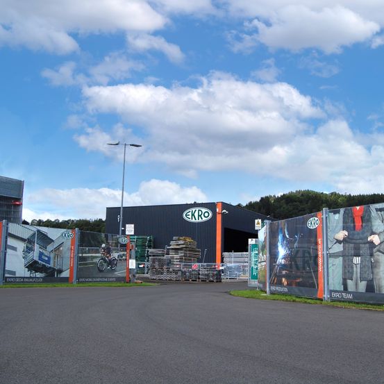 Das Äußere der Ekro-Fabrik mit Werbebannern, einem Radfahrer auf einem Plakat und einer Straßenlaterne im Vordergrund.