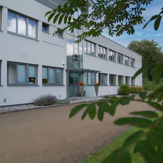 Ein modernes Bürogebäude mit Glasfenstern, zwei großen Vasen mit roten Blumen und einer Glastür mit dem Wort 'Ekro' darauf.