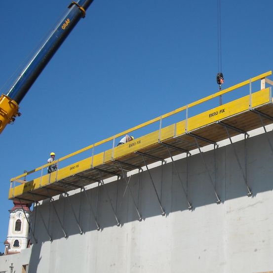 Baustelle mit einer Wand im Bau. Arbeiter in weißen Helmen und reflektierenden Westen stehen auf der Oberseite der Wand. Ein Kran befindet sich auf der linken Seite der Wand.