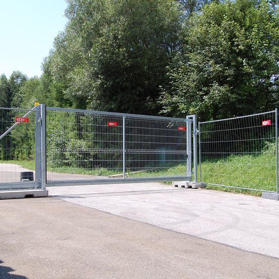 Ein Metalltor mit roten und weißen Schildern steht offen auf einer Straße, die in einen Wald führt.