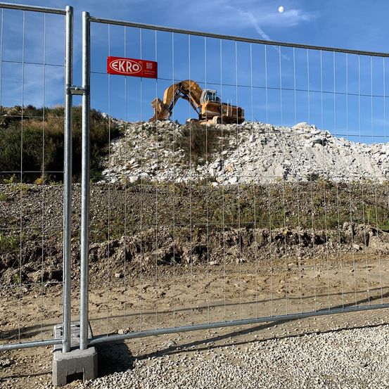 Baustelle mit einem Metallzaun und einem roten Schild mit der Aufschrift 'EKRO'. Ein gelber Bagger steht auf einem Haufen Steine.