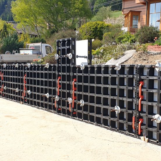 Eine lange Mauer aus ineinandergreifenden Betonblöcken mit orangefarbenen Griffen und Metallkappen wird vor einem Haus errichtet.