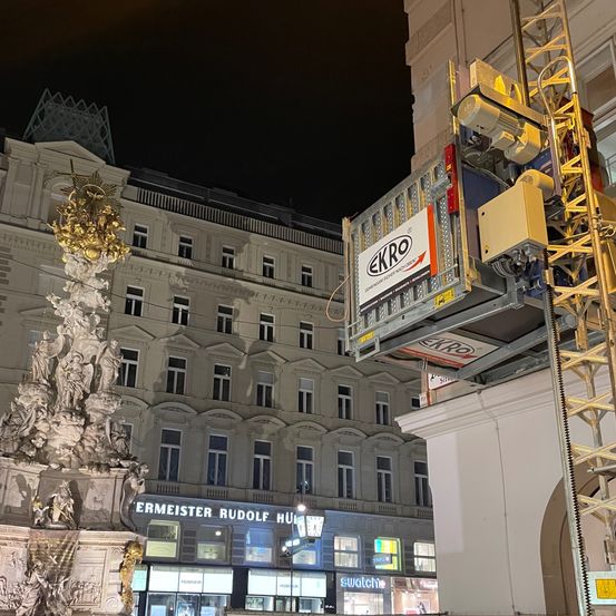 In der Nacht steht eine goldene Statue vor einem historischen Gebäude in Wien, Österreich. Ein hoher Kran mit einer gelben Plattform ist neben der Statue positioniert, wahrscheinlich für Wartungsarbeiten oder ein Ereignis.