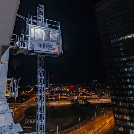 Ein großer silberner EKRO-Aufzug mit einem gelben Sicherheitsgeländer ist nachts an der Seite eines Gebäudes montiert. Die Stadt ist mit vielen Lichtern beleuchtet.
