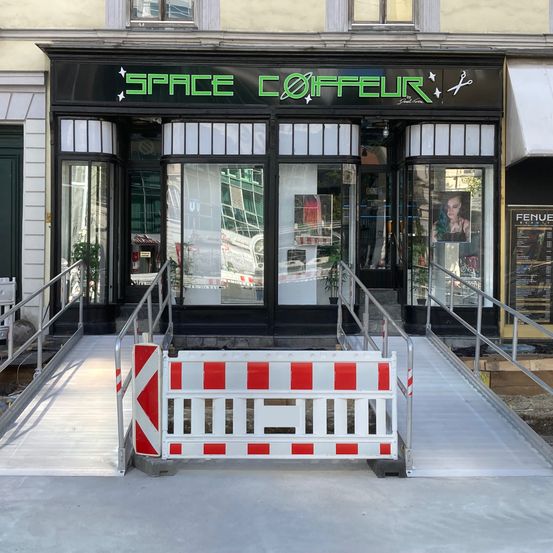 Vorderseite eines Ladens namens Space Coiffeur. Der Eingang hat eine Rampe für Rollstöhle mit einer Barriere auf dem Gehweg.