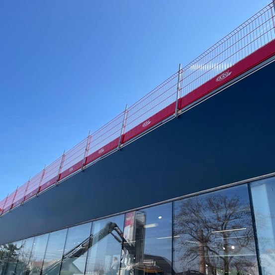 Die Außenseite eines Gebäudes wird umgebaut und zeigt ein rot gestrichenes Metallgeländer und eine blaue Wand. Das Gebäude hat große Glasfenster und eine Glastür.