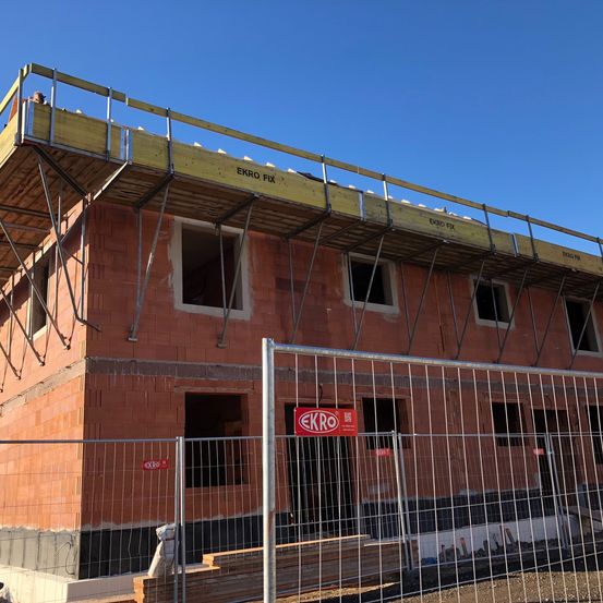 Ein Ziegelgebäude im Bau mit einem Metallgerüst und einer gelben Plattform auf dem Dach. Das Schild steht 'EKRO FIX'.