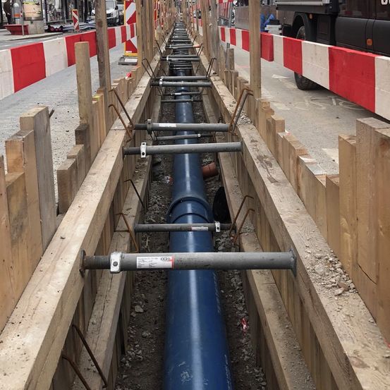 Eine Baustelle mit einer großen blauen Rohrleitung, umgeben von Holzbarrieren und Metallstangen, mit einem geparkten LKW in der Nähe.