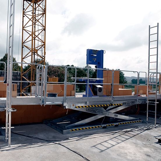 Ein Bauarbeiter mit weißer Schutzhelm geht auf einer Plattform mit Metallgittern. Auf der Plattform befinden sich mehrere braune Ziegelsteine, ein gelber Kran im Hintergrund und eine blaue Maschine.