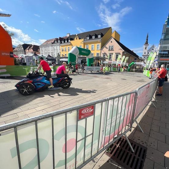 Eine Person auf einem blauen Motorrad fährt an einer Metallbarriere mit einem Banner vorbei, während eine Menschenmenge zusieht. Im Hintergrund gehen Menschen in der Nähe eines grünen Bogens.