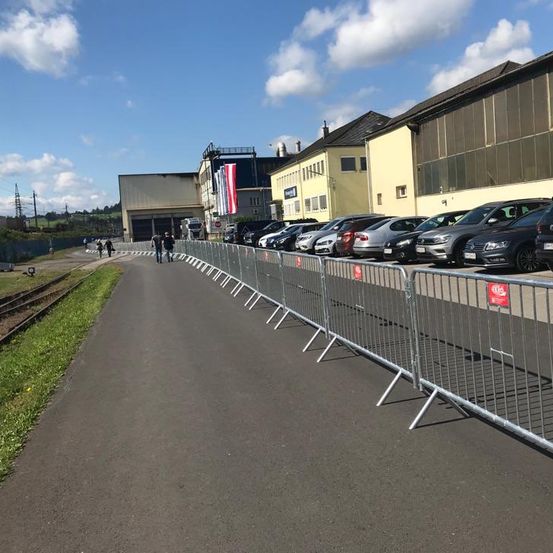 Eine Metallbarriere ist entlang einer Straße mit geparkten Autos aufgestellt, in der Nähe eines Gebäudes mit Flaggen und einem klaren blauen Himmel mit Wolken.