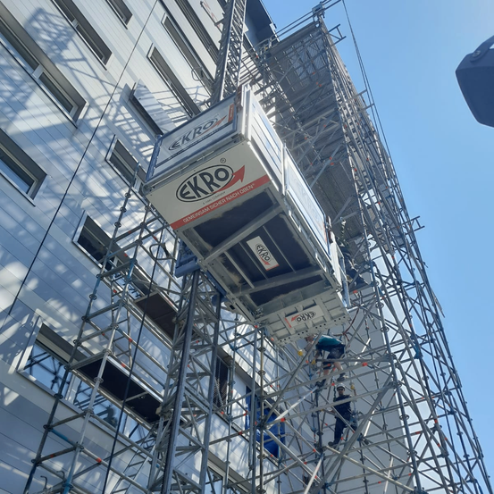 Ein Gebäude im Bau mit Gerüsten und Arbeitern. Ein großer weißer Ekro Bauaufzug ist am Gerüst montiert.