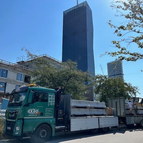Ein grüner Lkw ist auf der Straße geparkt, im Hintergrund befindet sich eine Baustelle mit einem hohen Gebäude im Bau, einem Kran und anderen Gebäuden.